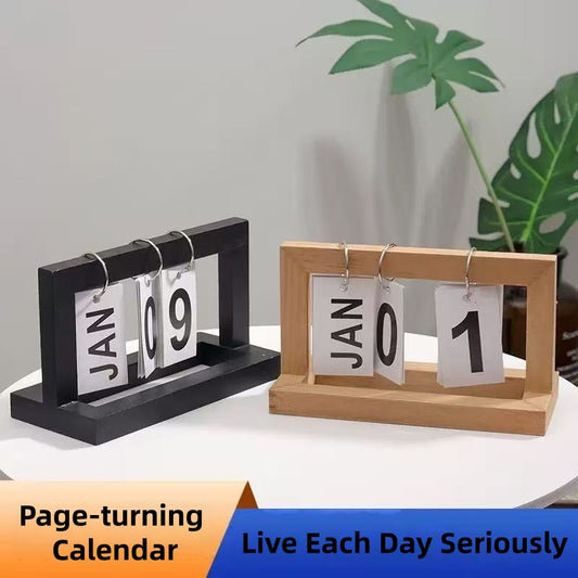 Two page-turning calendars, one black and one wooden, on a white surface with a plant in the background.